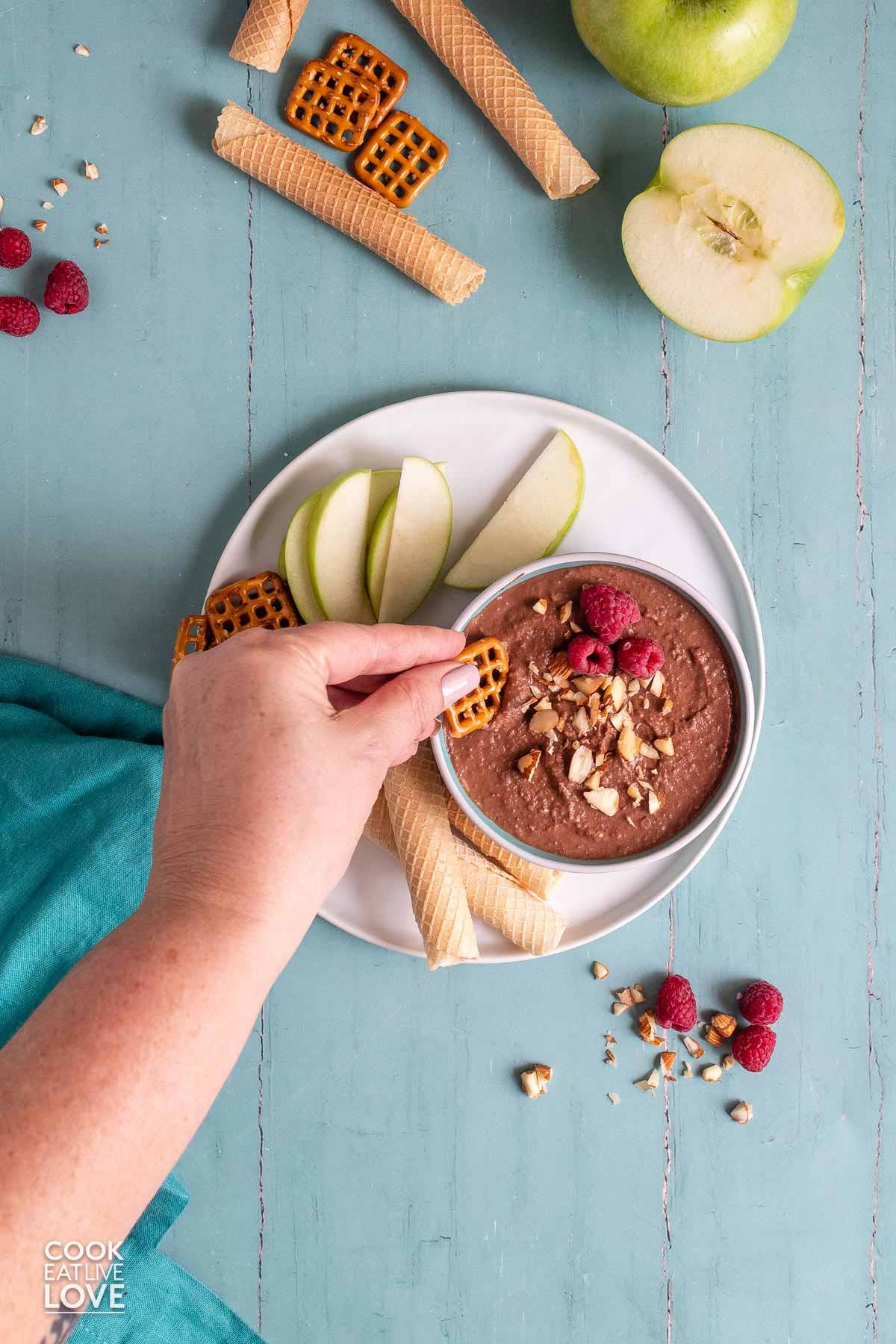 A bowl of dark chocolate hummus on the table with apple slices, pretzels and cookies and a hand dipping in a pretzel.