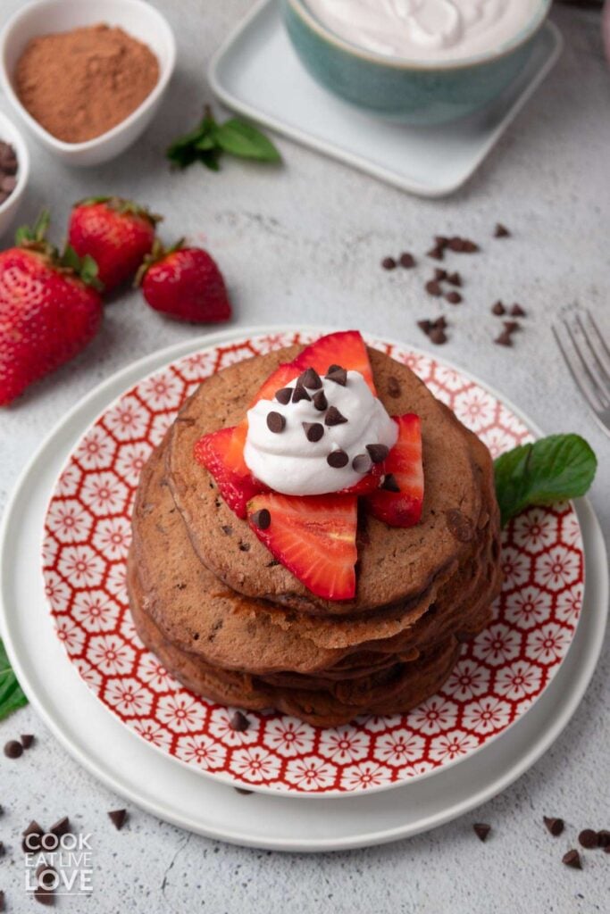 A stack of chocolate protein pancakes on a plate garnished with strawberry slices and whipped cream.