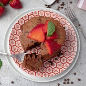 A stack of chocolate protein pancakes on a plate topped with sliced strawberries and a bite on a fork.