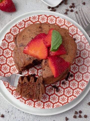 A stack of chocolate protein pancakes on a plate topped with sliced strawberries and a bite on a fork.