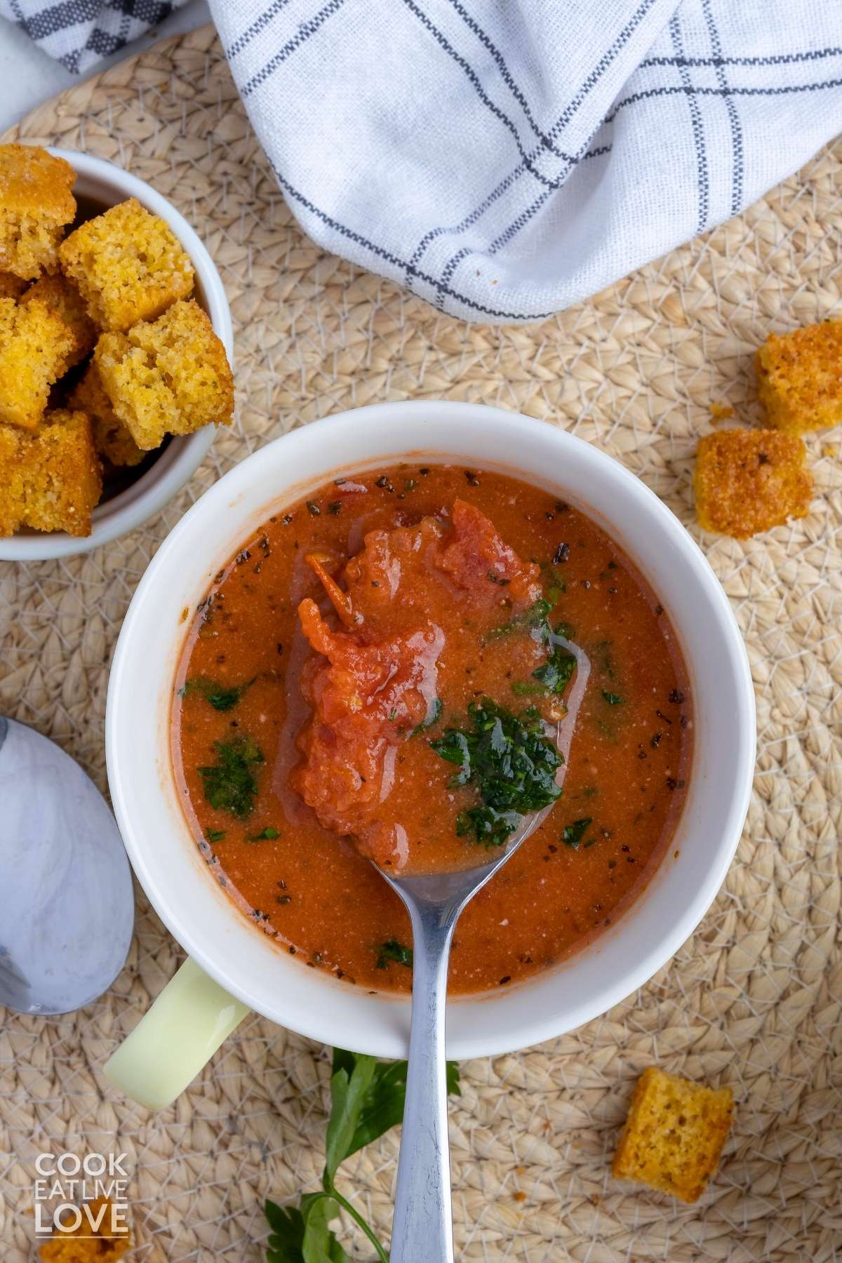 A bowl of dairy free tomato soup with a spoon held over the bowl with a bit of soup and a bowl of croutons on the side.