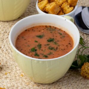 A cup of dairy free tomato soup on the table with some croutons and another cup of soup in the background.