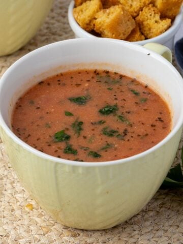 A cup of dairy free tomato soup on the table with some croutons and another cup of soup in the background.