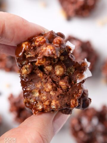 A hand holding up a dark chocolate quinoa crisp with a bite taken to show the inside.
