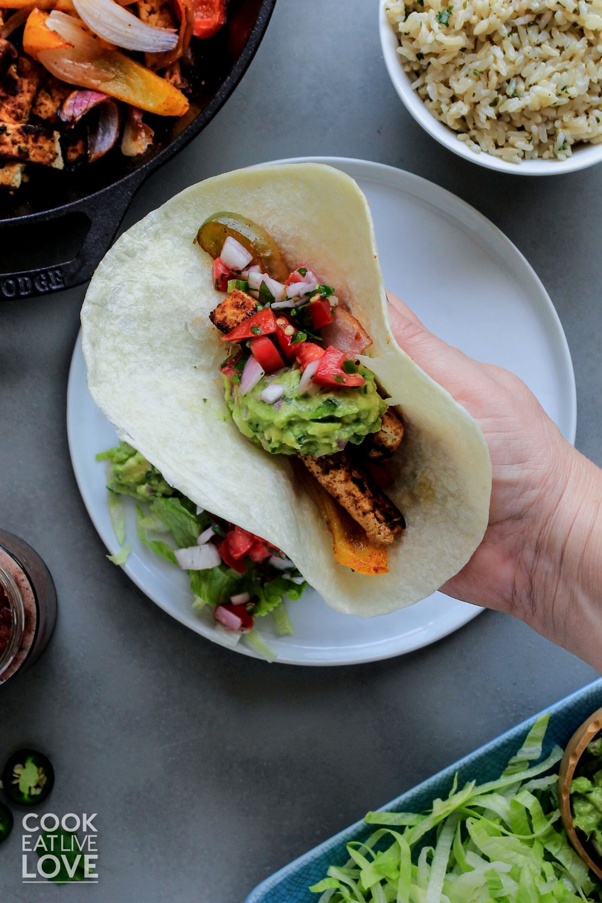 A hand holding a fajita up over a plate with various toppings and sides for fajitas around it