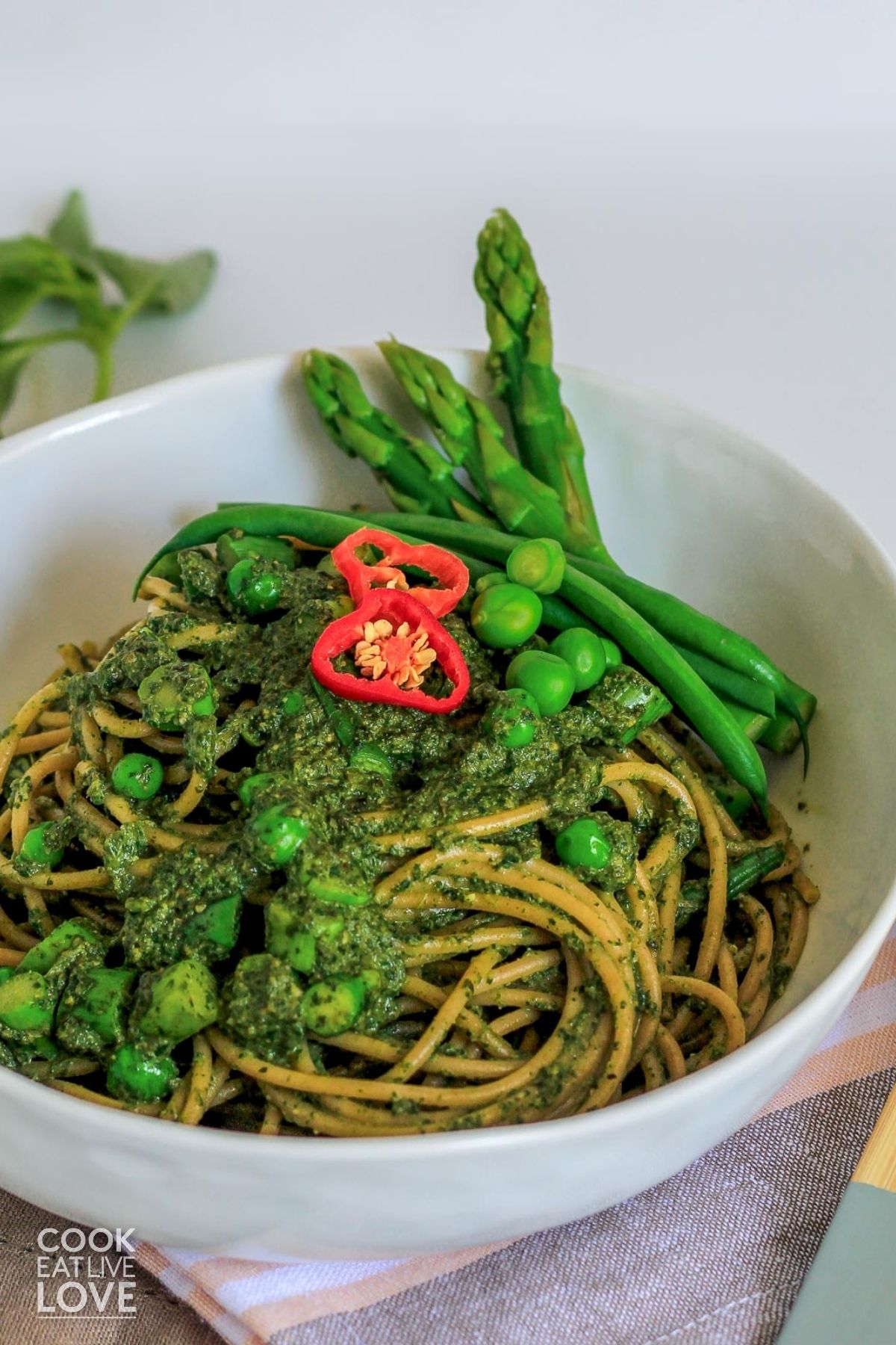 Green spaghetti in a bowl with peas, asparagus, and red chilies on top.