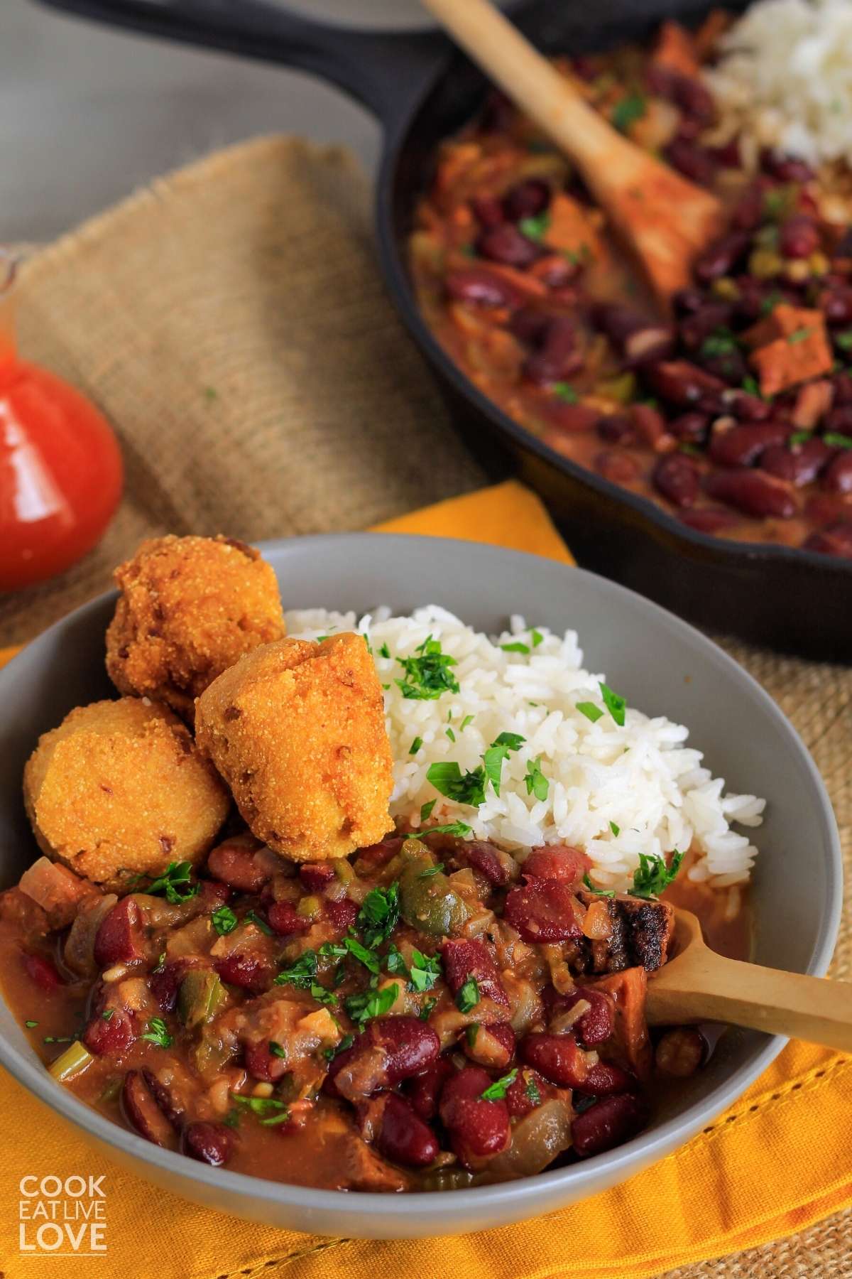 A bowl of vegan red beans and rice on the table with a skillet with more in the background and hush puppies in the bowl.