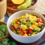 A bowl of 5 ingredient mango salsa on the table with fresh produce around it.