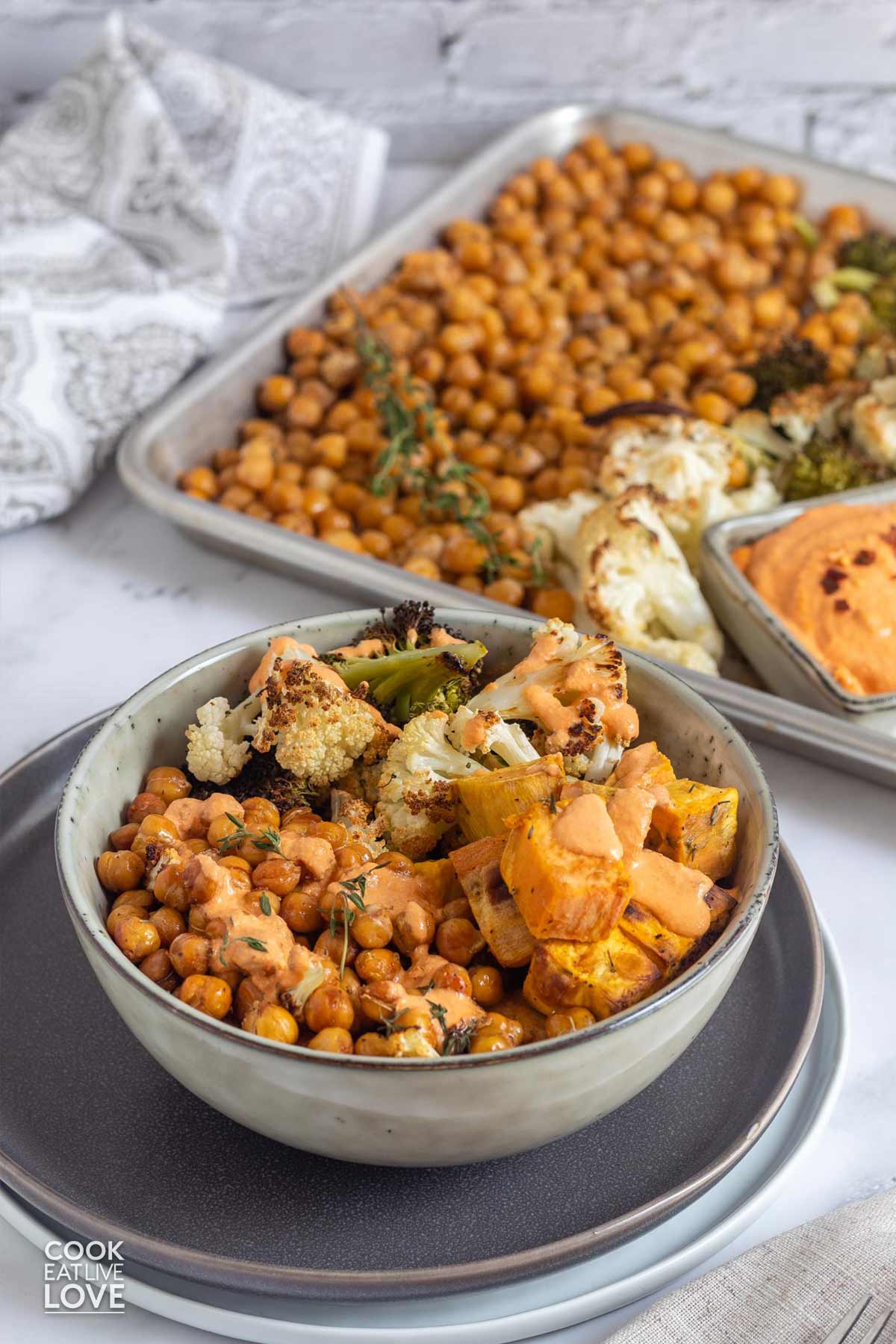 A bowl of mediterranean roasted vegetables in a bowl on the table with sheet pan of veggies behind it.