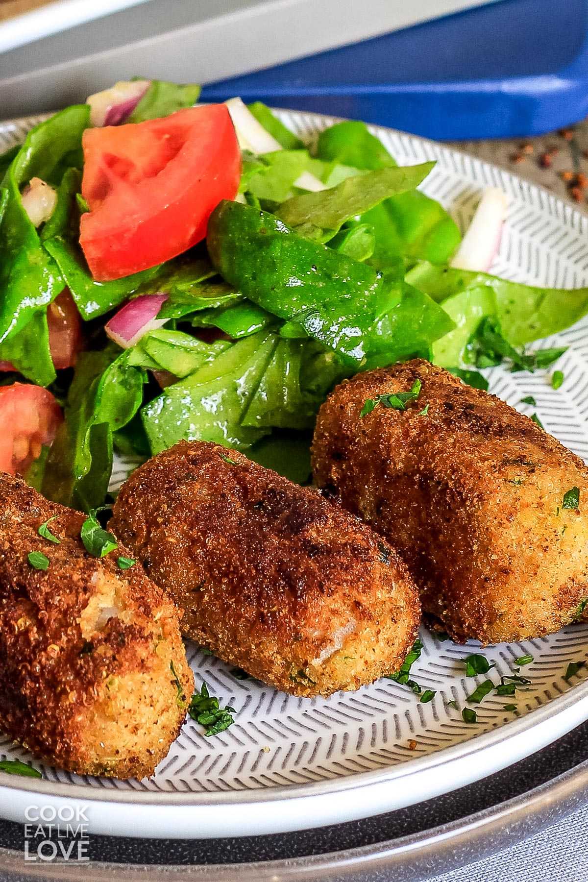Cooked potato croquettes on plate ready to eat with a salad in the background of the plate.