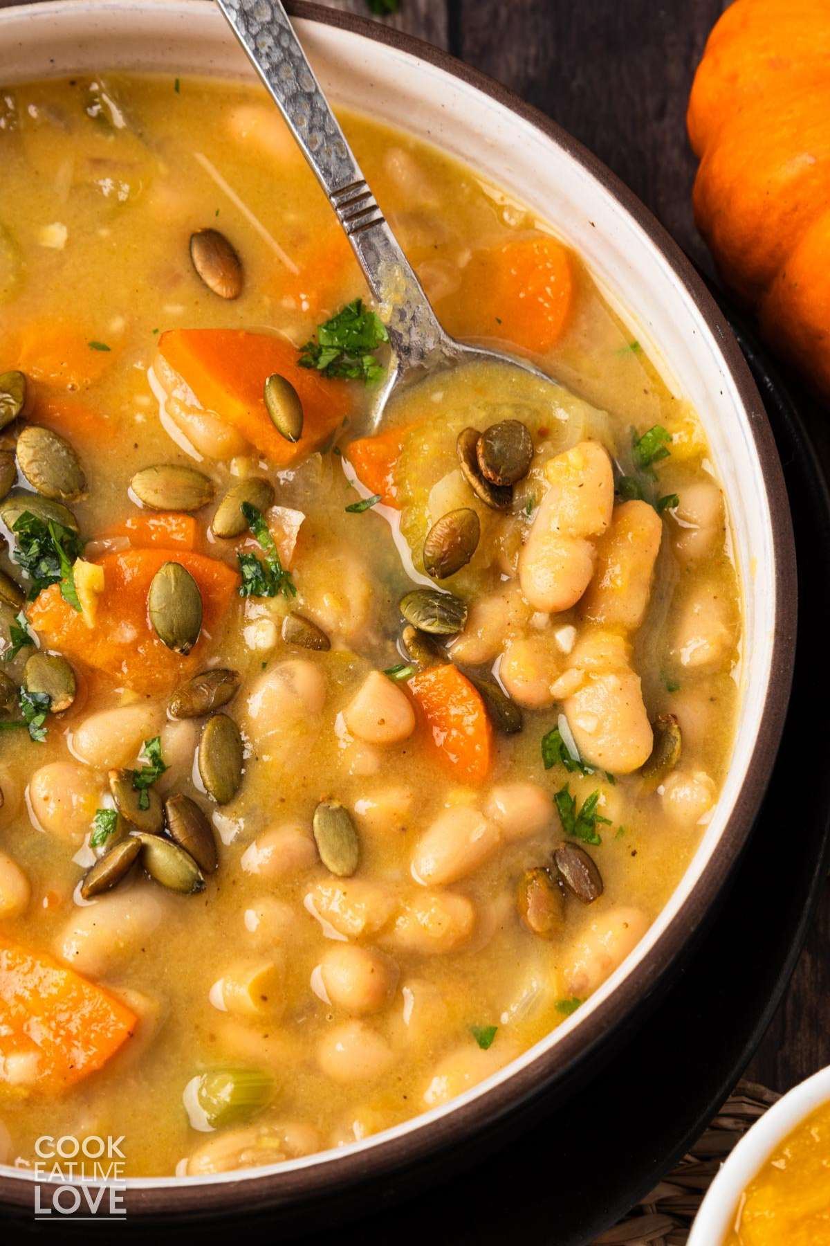 A bowl of pumpkin white bean soup with a spoon in the bowl and parsley and pumpkin seeds on top.