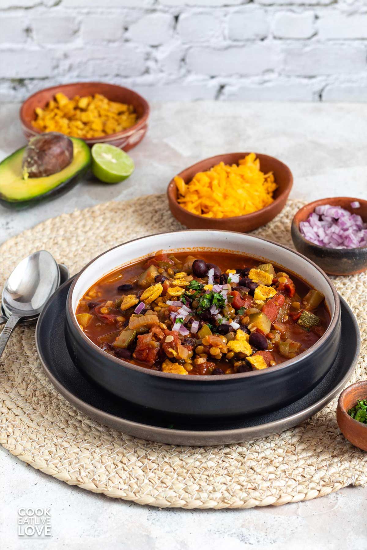 A bowl of vegetarian chili on the table with bowls of cheese, red onions and cilantro.