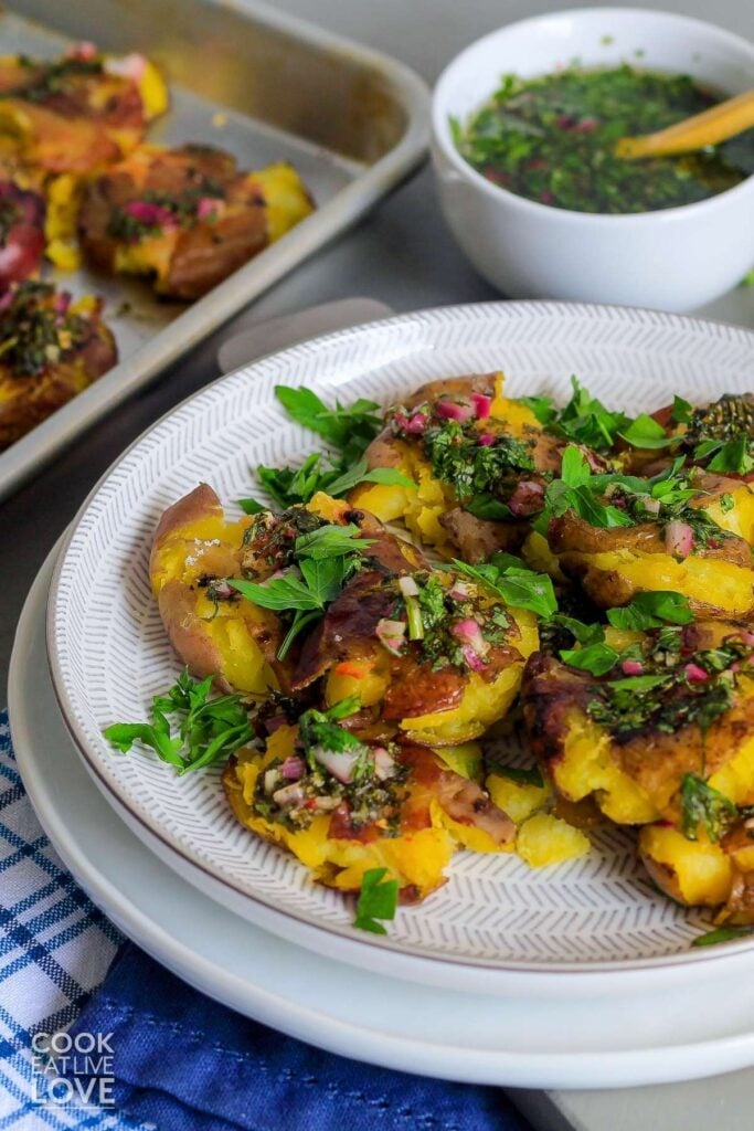 Smashed red potatoes on a white plate with more on a baking pan and a small bowl of chimichurri in the background.