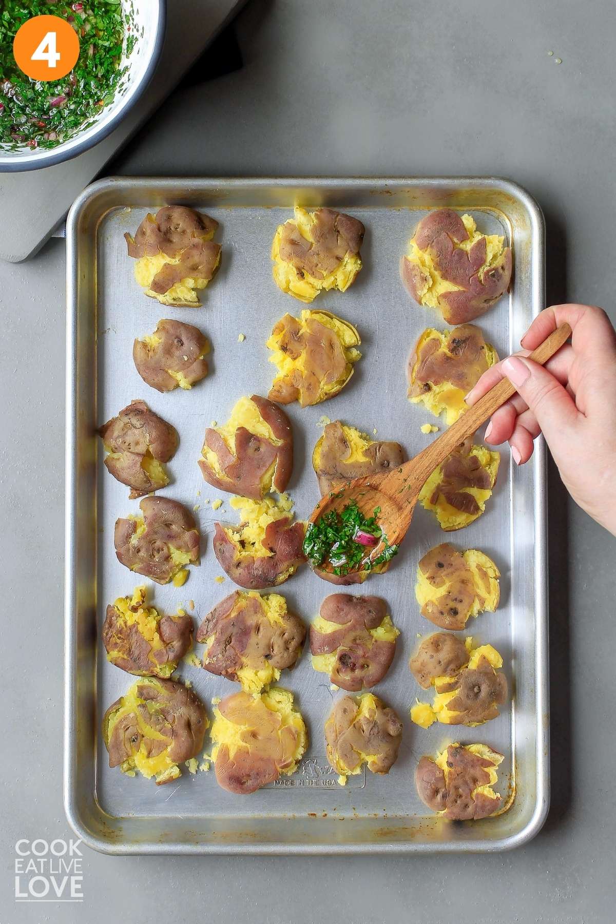 Smashed red potatoes on a pan with chimichurri sauce in a spoon adding to the top of the potatoes.