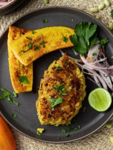 A tacu tacu on a brown plate with two plantains, onion salad, and a lime.