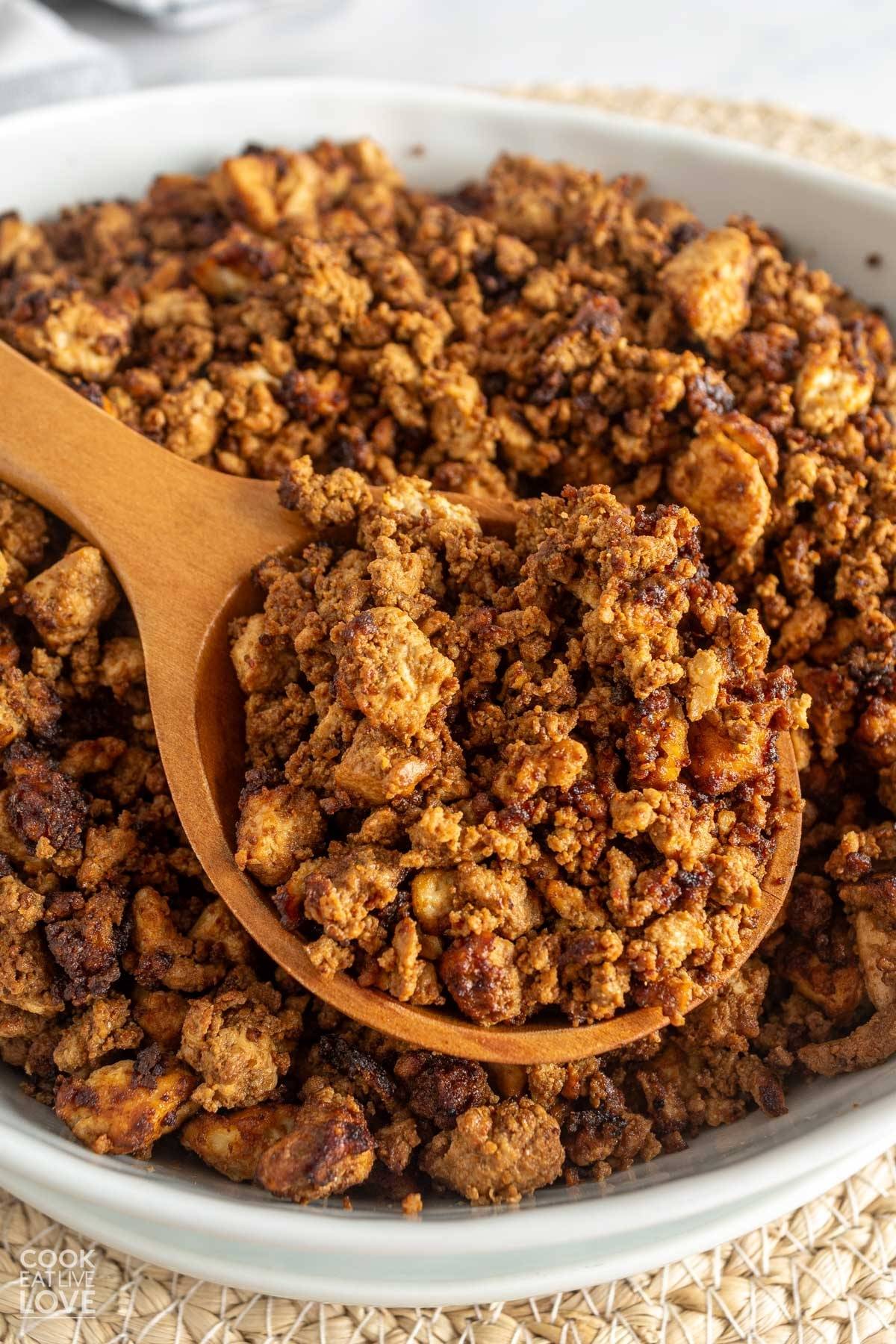 Cooked tofu ground beef in a bowl with a wooden spoon lifting some up.