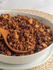 A bowl of tofu ground beef crumbles on the table with a wooden spoon in the bowl and some crumbles on it.