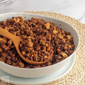 A bowl of tofu ground beef crumbles on the table with a wooden spoon in the bowl and some crumbles on it.