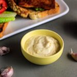 Vegan roasted garlic aioli in small bowl with garlic cloves and small wooden spoon to side.