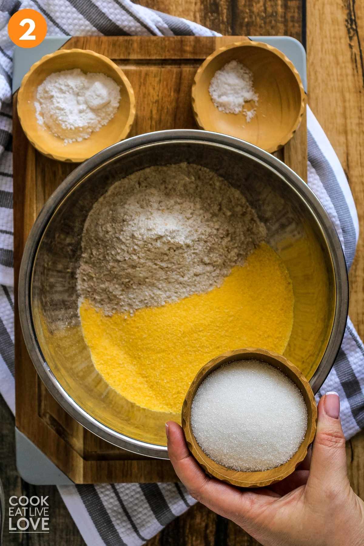 Dry ingredients such as corn meal and flour are in a mixing bowl, a hand is holding up the sugar to add and other ingredients are shown to the side.