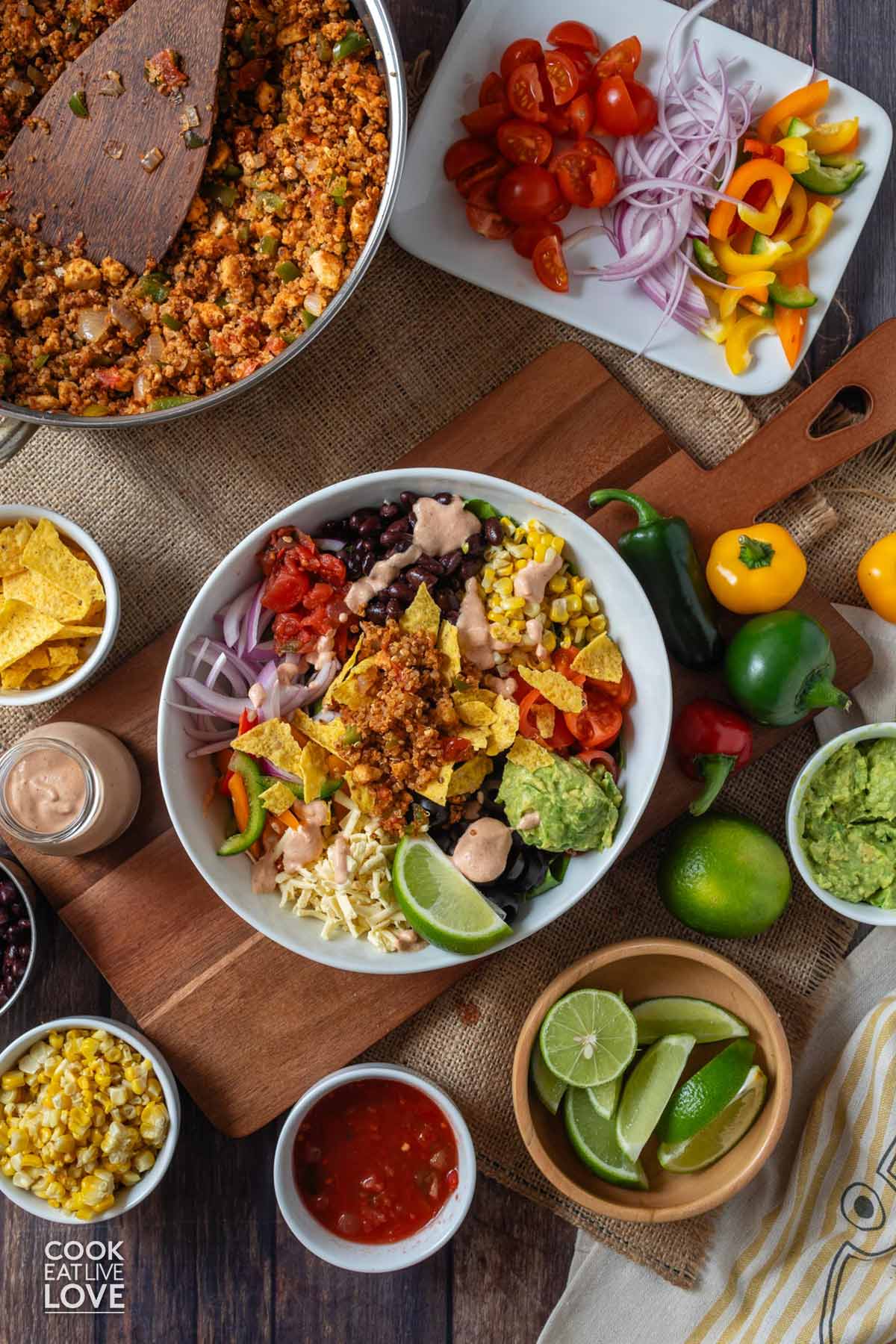 A vegan taco salad on the table surrounded by a plate with the toppings, bowls of avocado, chips, and a skillet of the tofu taco meat.