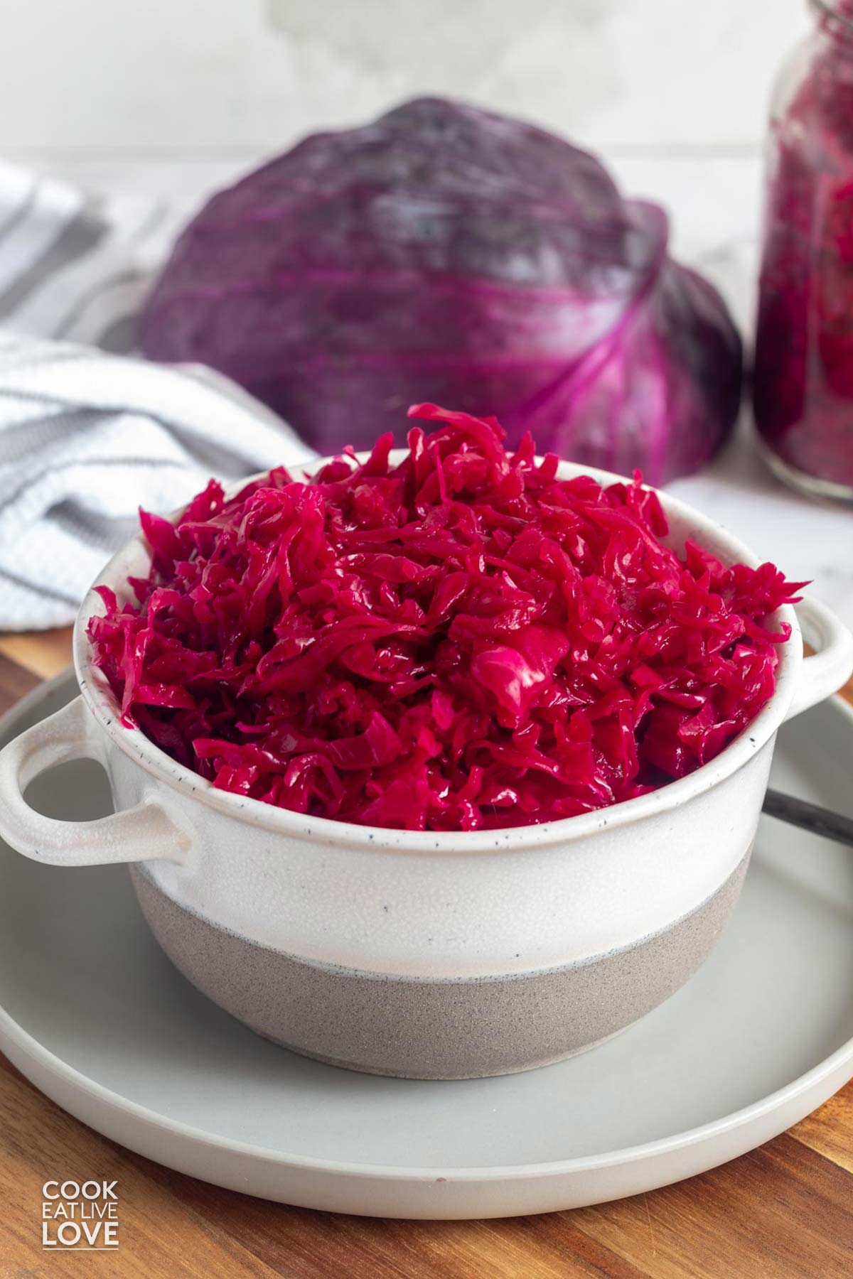 A bowl of red cabbage sauerkraut on a plate with purple cabbage in the background and a jar of more sauerkraut.