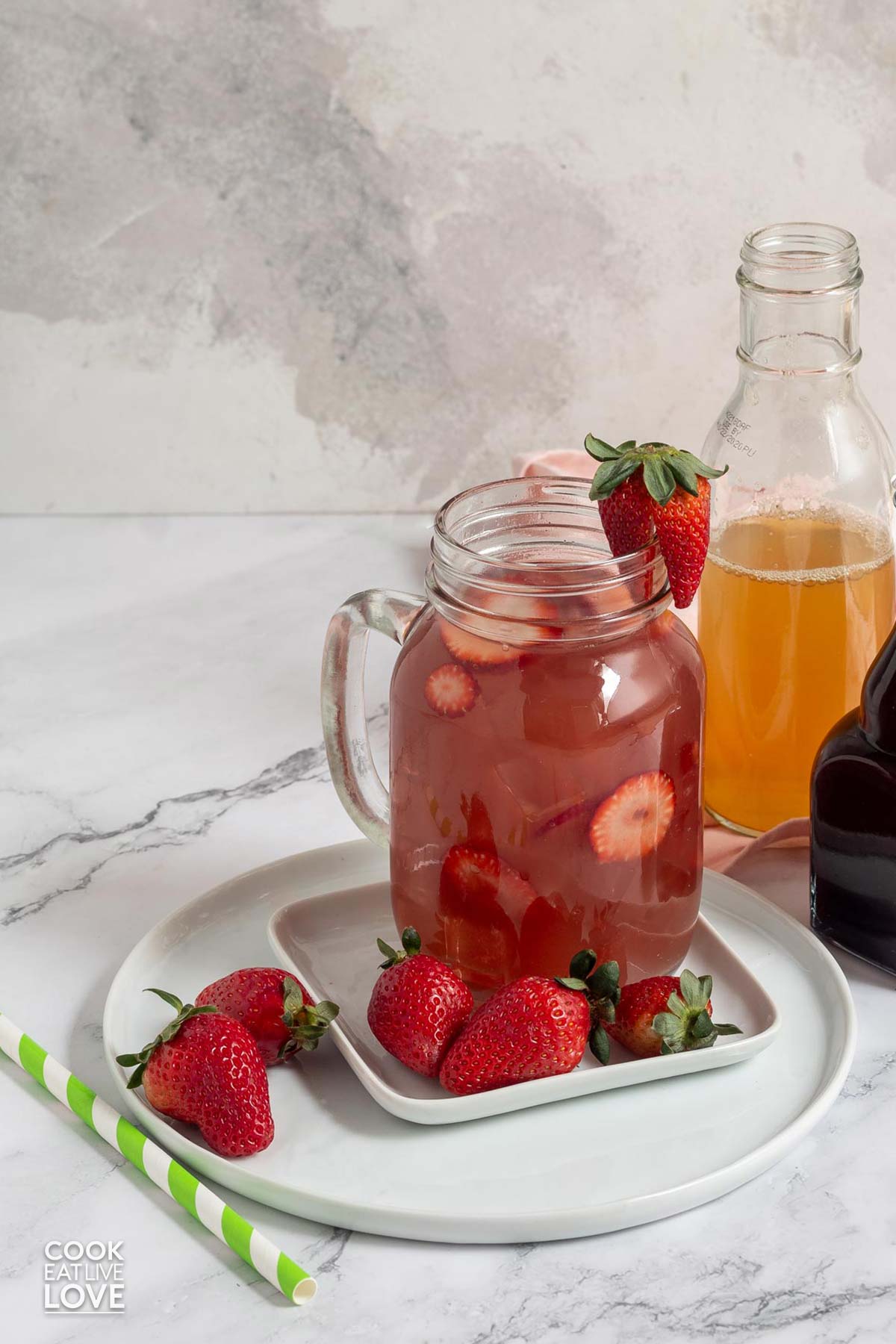 A glass of strawberry acai refresher on a plate with fresh strawberries and jar of syrup and green tea in the back.
