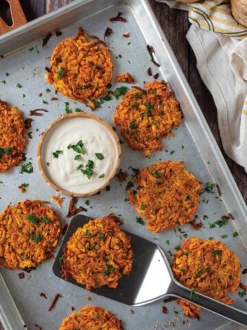 Sweet potato hashbrowns on a baking tray with a spatula underneath one and a bowl of sauce in the middle.