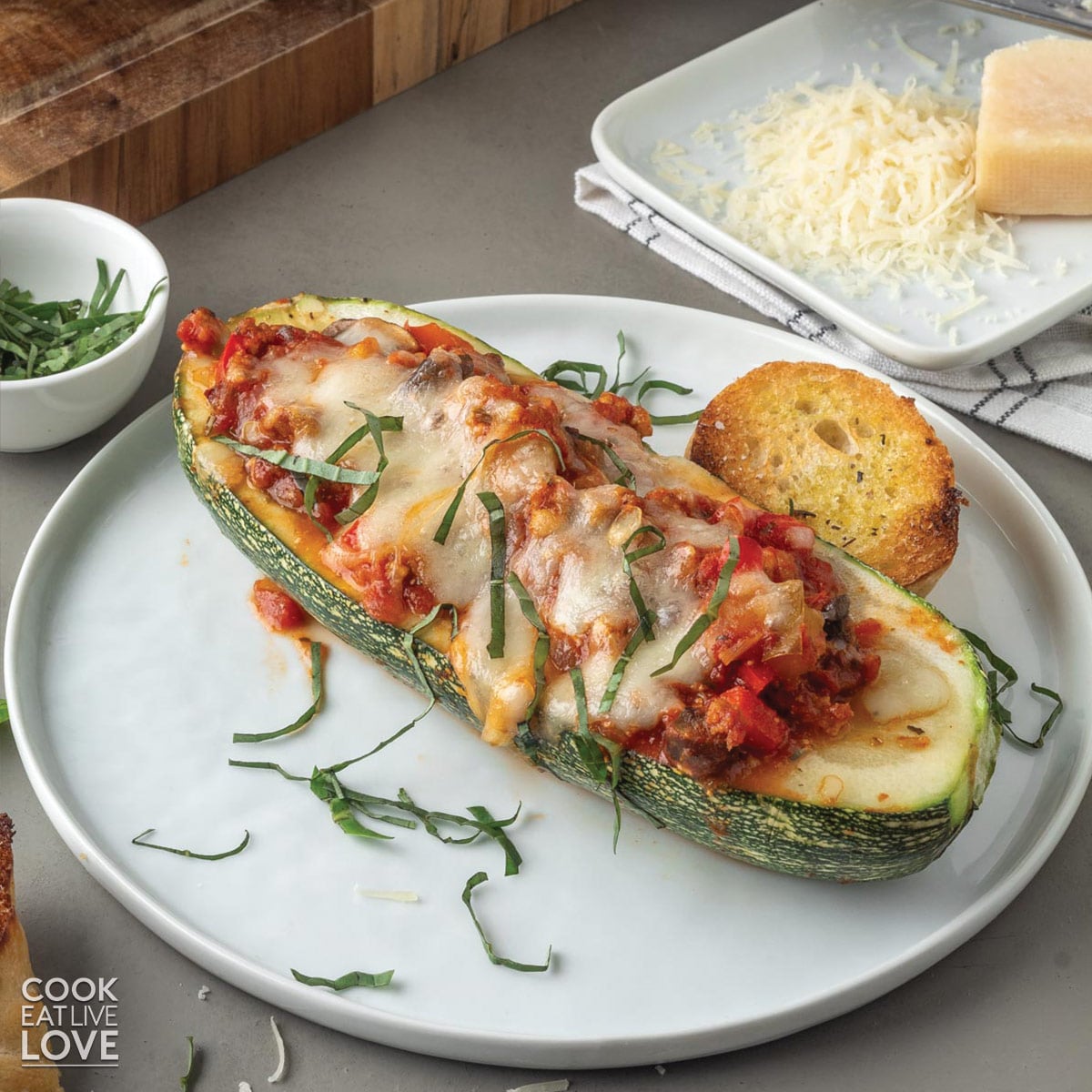 Stuffed vegetarian zucchini on a plate garnished with fresh basil and a plate of grated cheese in the back.