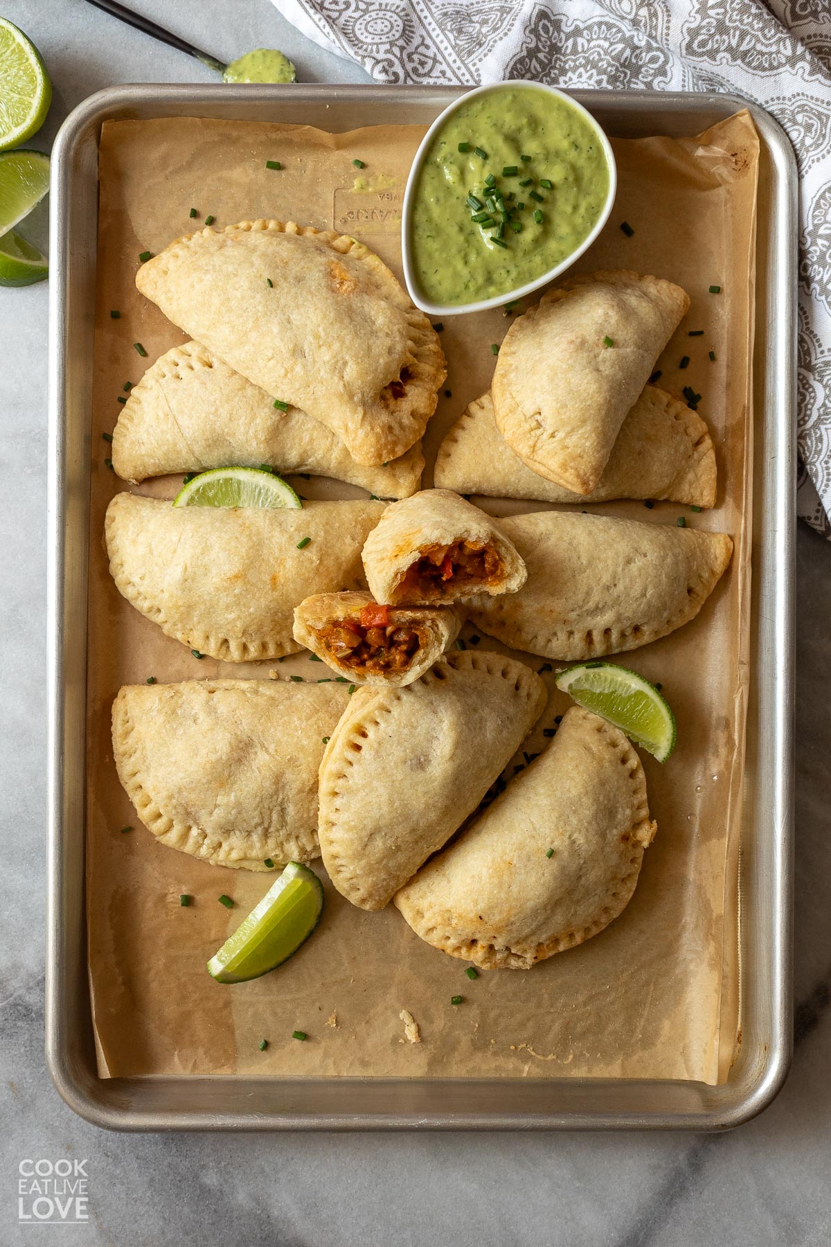 Baked empanadas on a baking tray with a bowl of green sauce and one in the middle broken in half.