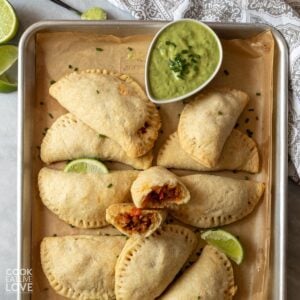 Vegean empanadas on a baking tray with a bowl of green sauce and one in the middle broken in half.
