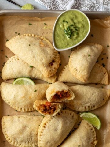 Vegean empanadas on a baking tray with a bowl of green sauce and one in the middle broken in half.