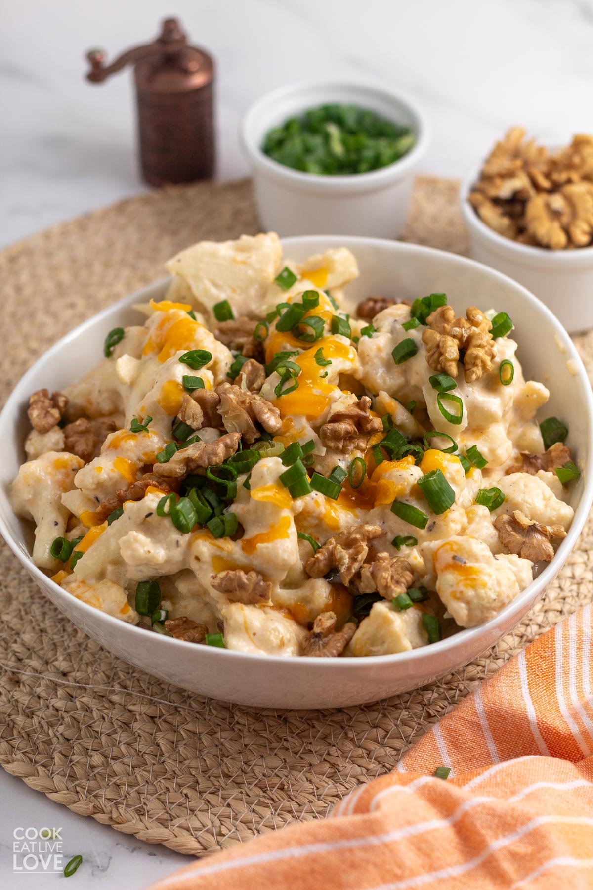 crockpot cauliflower casserole garnished with walnuts and green onions in a white bowl with small bowls of the garnishes behind it.