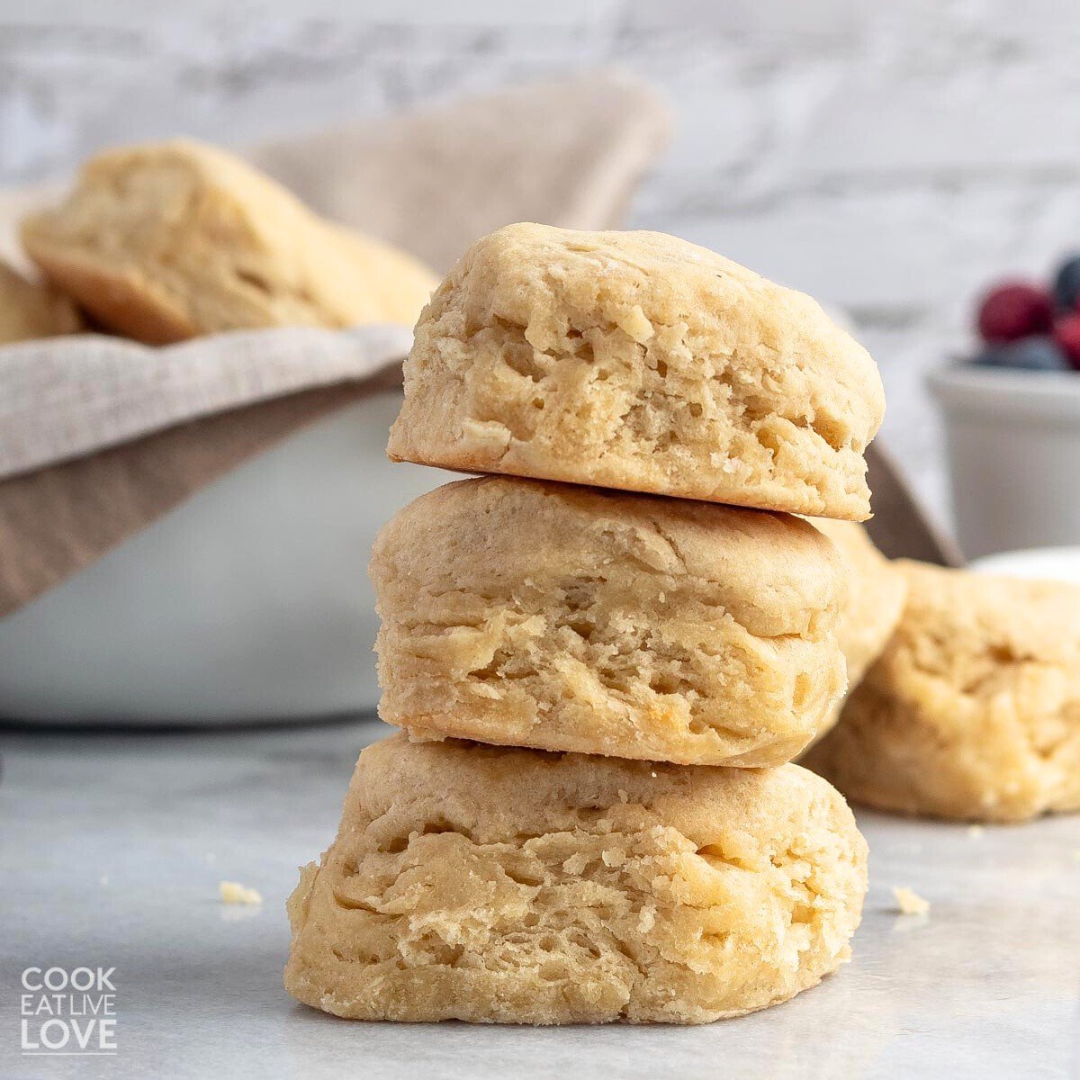 front view of three dairy free biscuits stacked on top of each other on a marble surface. with more biscuits and berries in the background.