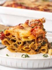 A serving of meatless baked ziti served up on a plate with casserole dish in the background.
