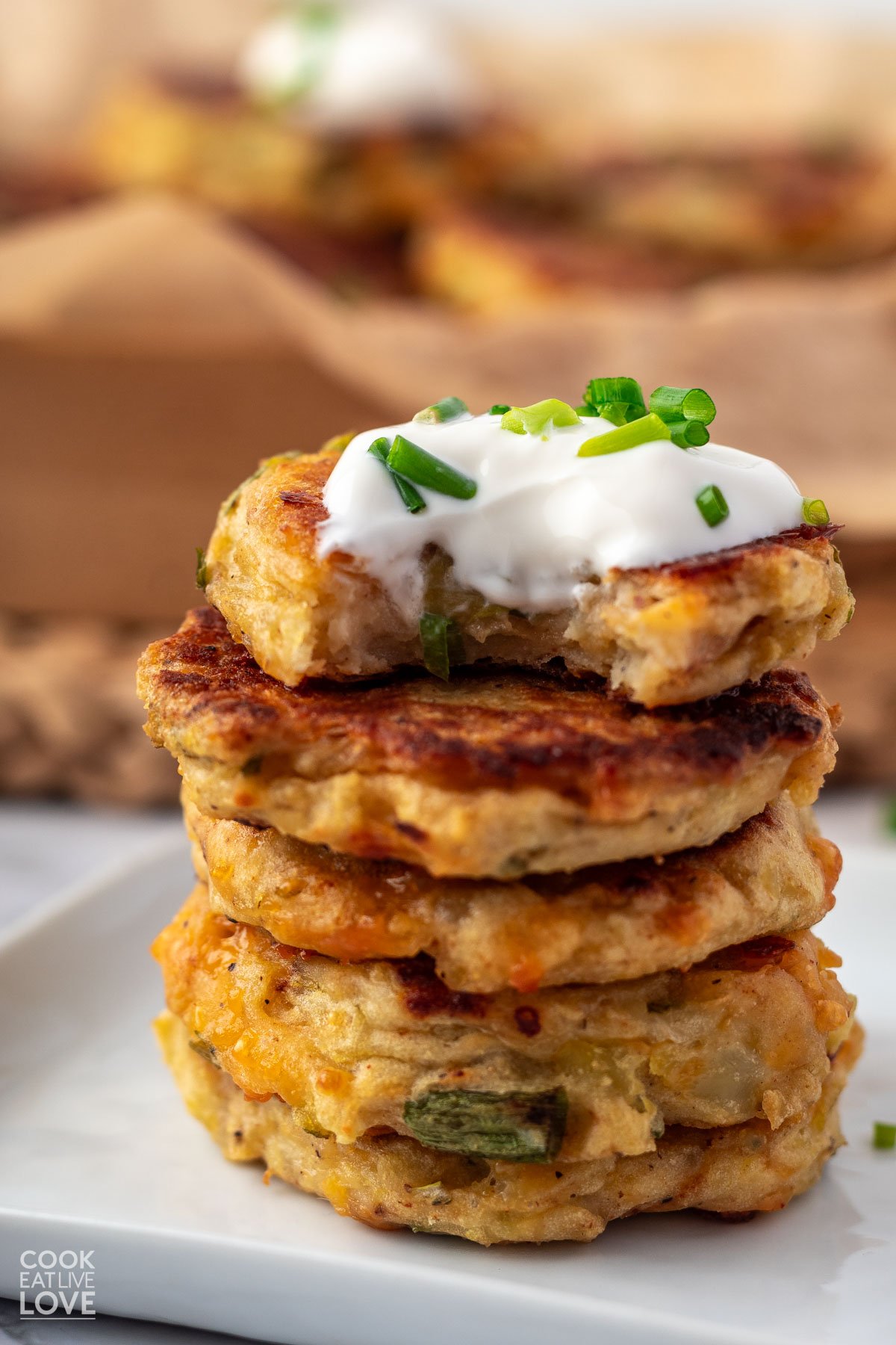 Stack of 5 potato fritters on a white plate with the top one missing a bite to show the inside and topped with sour cream and chives.