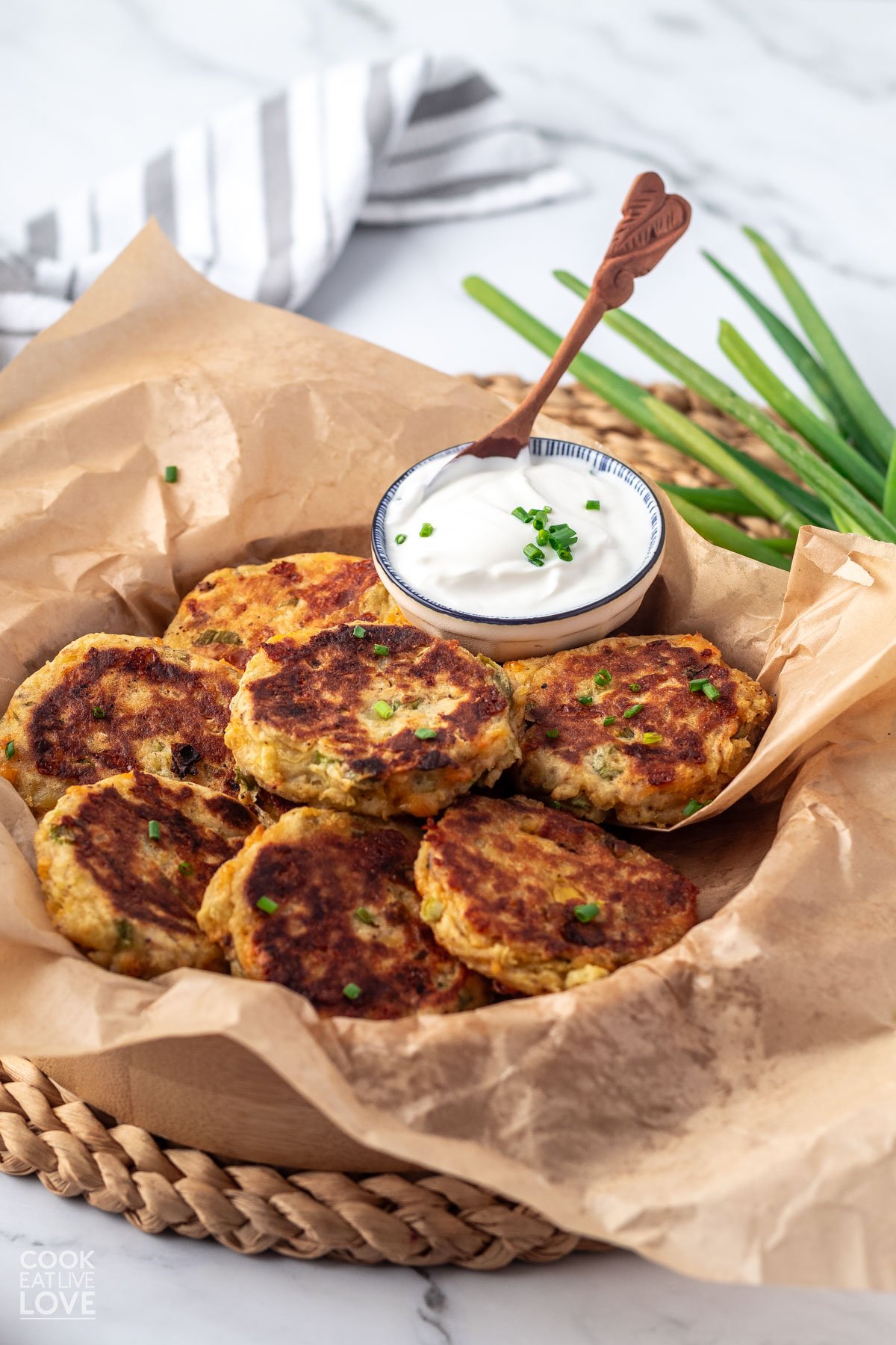 Potato fritters on parchment paper in a basket garnished with chives and a small dish of sour cream with a spoon in it.