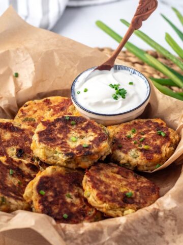 close-up of potato fritters on parchment paper in a basket and a small dish of sour cream with a spoon in it.