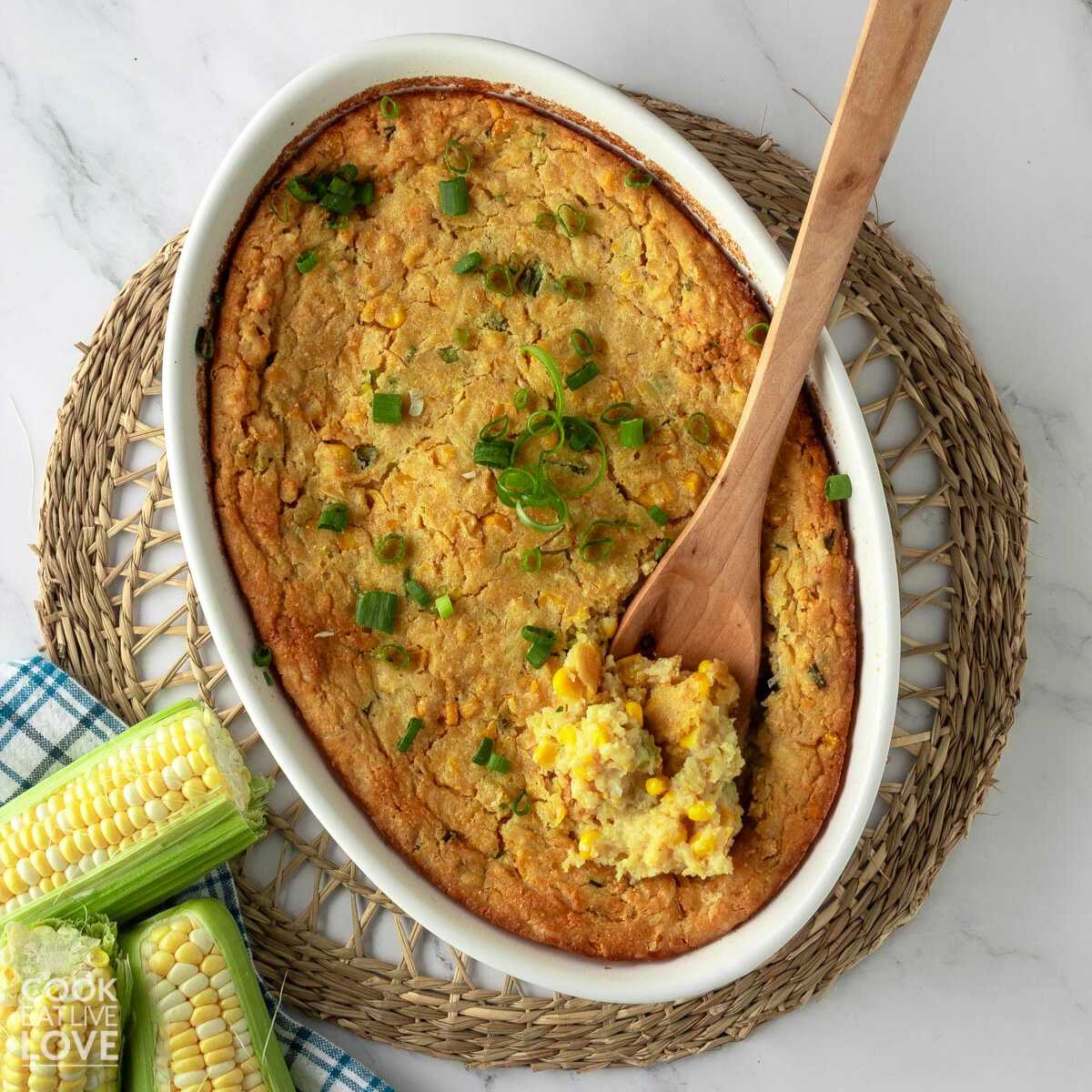 Close-up overhead shot of vegan corn casserole in a white baking dish with a wooden spoon in it scooping some of it out of the dish.