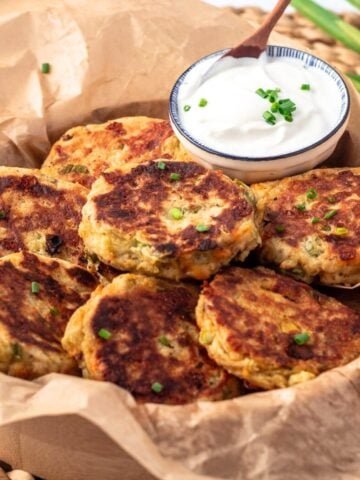 Potato fritters on parchment paper in a basket and a small dish of sour cream with a spoon in it.