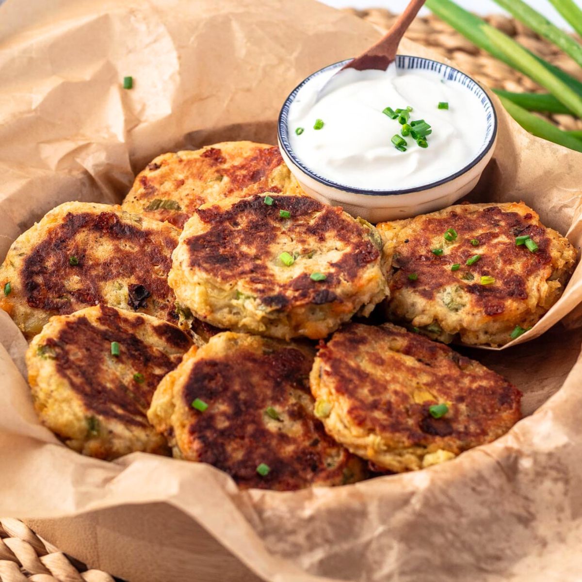 Potato fritters on parchment paper in a basket and a small dish of sour cream with a spoon in it.