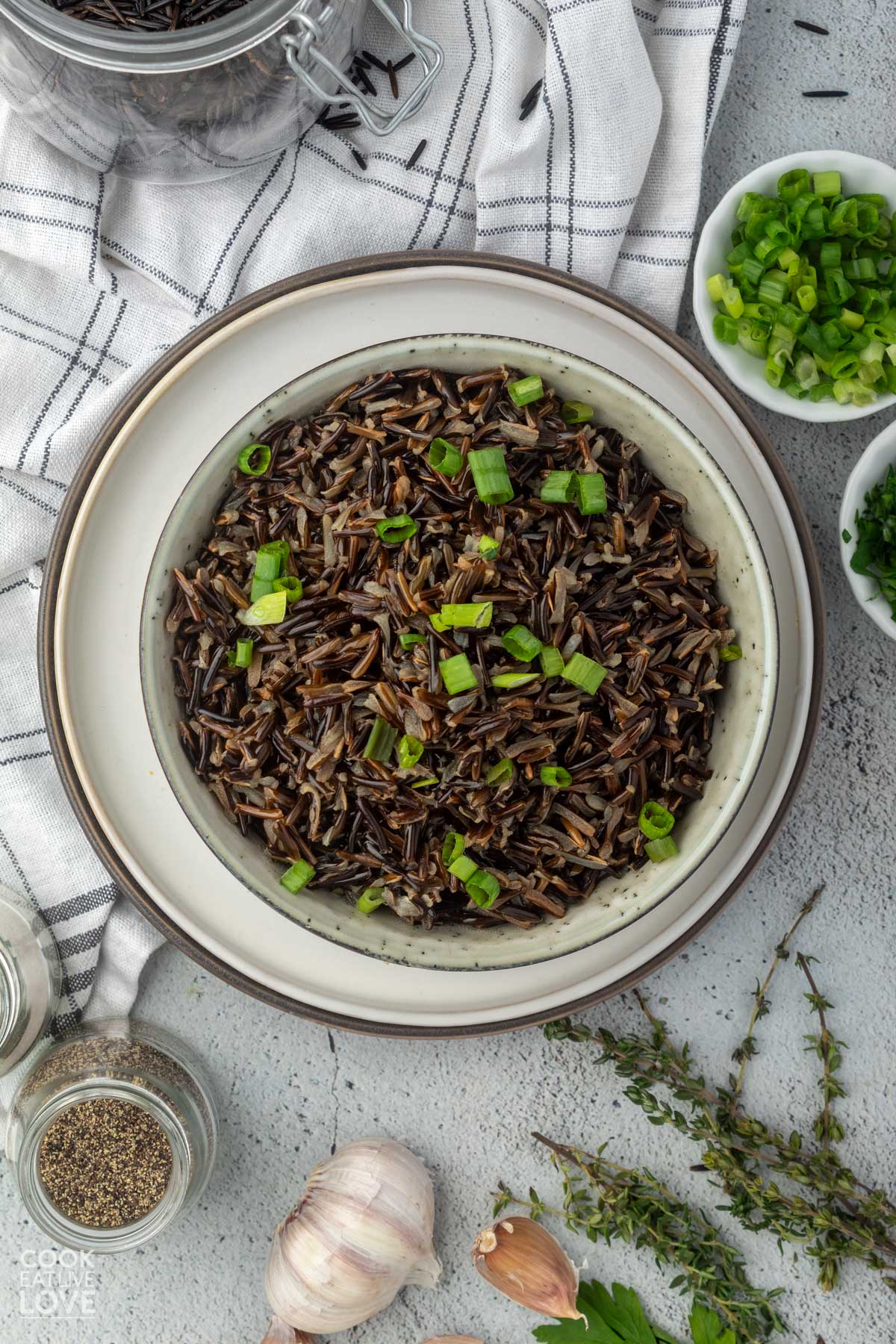 Wild rice in a bowl set on a plate on the table with little bowls of green onions and fresh herbs.