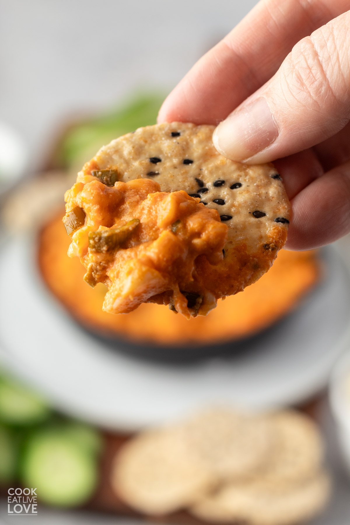 A hand holding up a cracker with some vegan buffalo chicken dip on it up over the table.