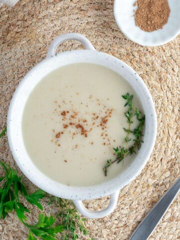 Vegan bechamel sauce in white bowl on the table with a spoon to the side and a bowl of nutmeg.