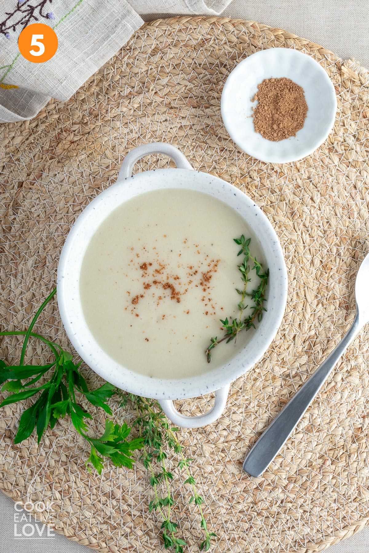 Vegan bechamel sauce served up in a small bowl on the table with fresh thyme on top and some parsley on the table.