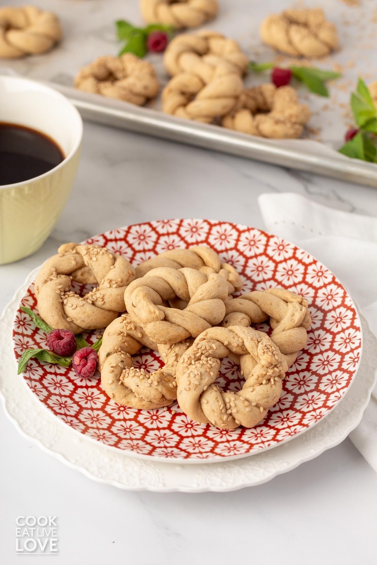 A plate of anise cookies with mint and berries and a cup of coffee and baking tray in the background.