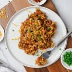 A white plate of vegetarian goulash with a bite on a fork to the side and bowls of red pepper flakes and green onions.
