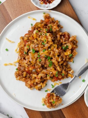 A white plate of vegetarian goulash with a bite on a fork to the side and bowls of red pepper flakes and green onions.