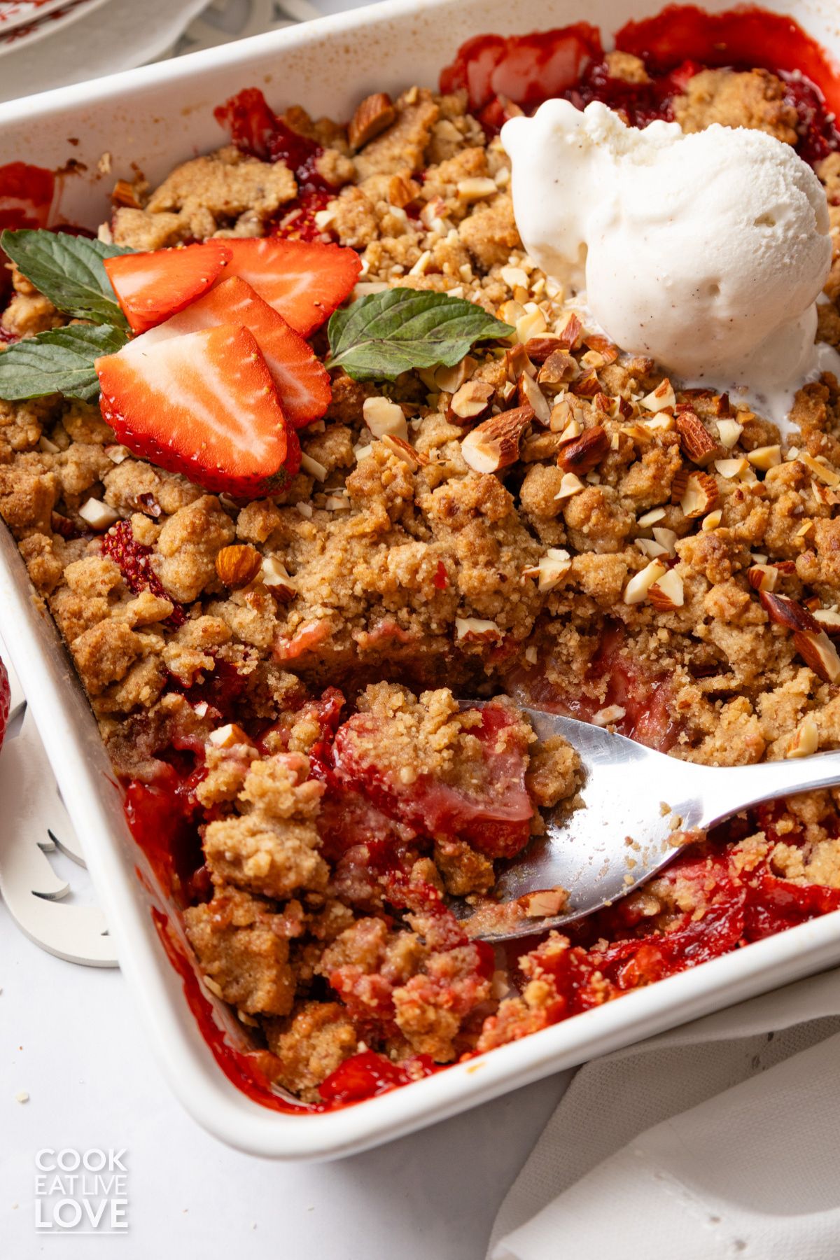 Almond strawberry crumble in a white baking dish with a spoon resting in the corner.
