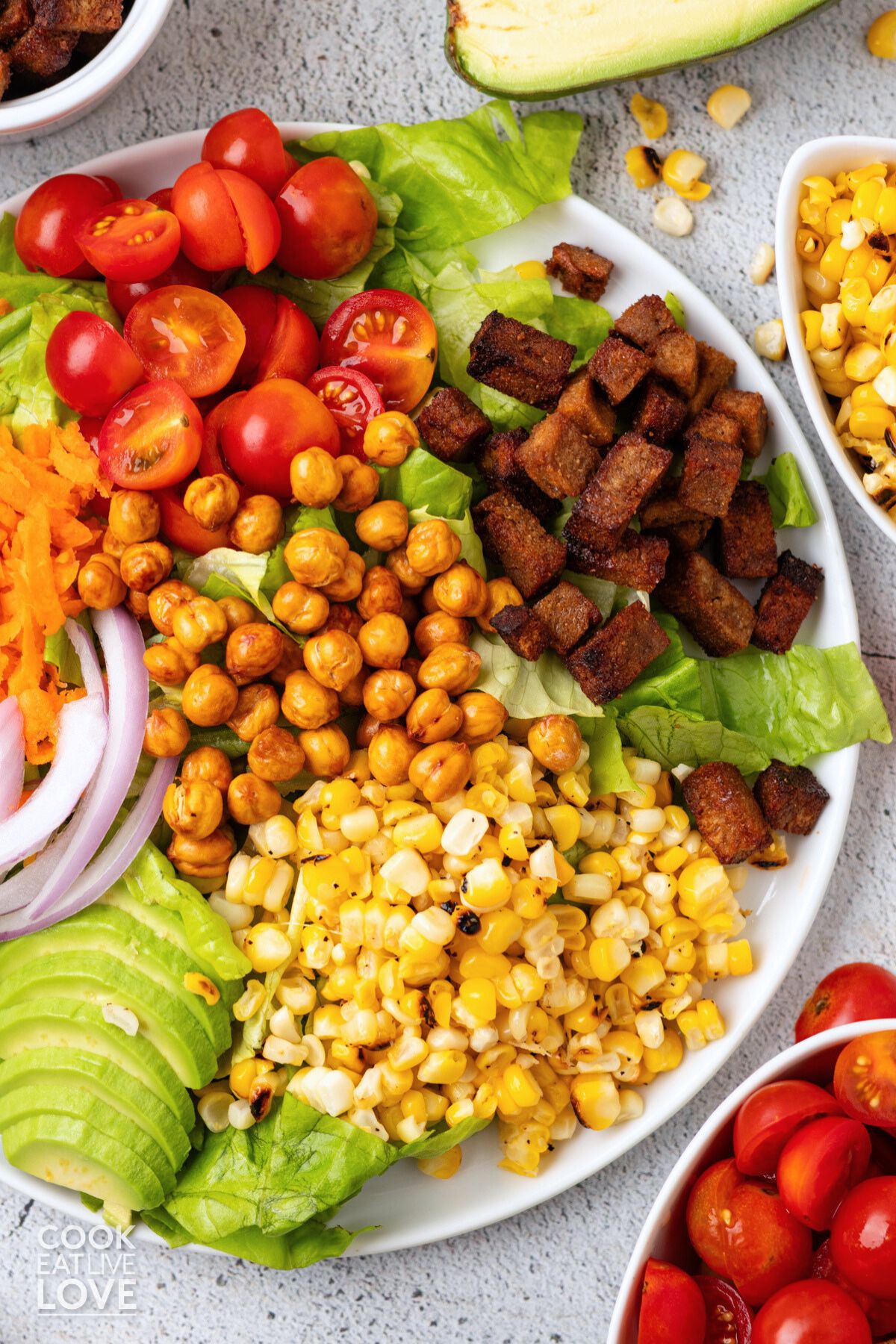 Tomatoes, tofu bacon, corn, avocado, chickpeas, and red onion on lettuce on a plate.